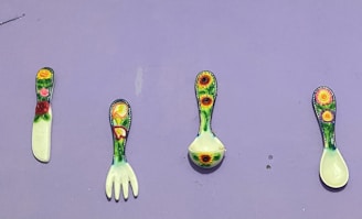 Four colorful utensils are arranged against a light purple background. Each utensil features a different floral pattern with vibrant yellows, greens, and reds. The utensils include a knife, a fork, a ladle, and a spoon.