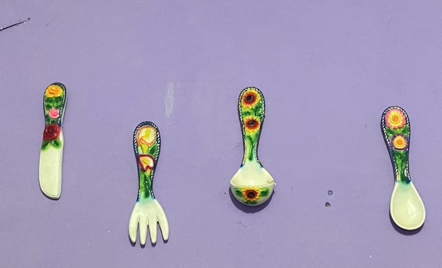 Four colorful utensils are arranged against a light purple background. Each utensil features a different floral pattern with vibrant yellows, greens, and reds. The utensils include a knife, a fork, a ladle, and a spoon.