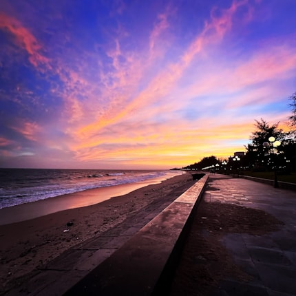 A vibrant sunset over a beach with dramatic clouds in shades of pink, orange, and purple. The shoreline features gentle waves and a smooth sandy surface. A long promenade runs parallel to the beach, lined with lit street lamps and trees in silhouette.