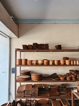 A set of wooden shelves displays a variety of neatly stacked wooden kitchenware, including bowls, plates, trays, and cutlery. The items are carefully organized, and the shelves are placed against a light-colored wall, adjacent to a window that lets in natural light.