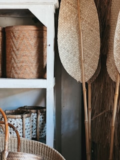 Several woven baskets are arranged on a white wooden shelf. The baskets display intricate patterns and are made from natural materials. Tall woven fans or paddles lean against the wall, complementing the earthy tones of the scene.