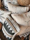 A collection of decorative pillows featuring intricate patterns and textures, some with black and white geometric designs and others with woven and embroidered details. The pillows are stacked closely together, creating a cozy and artisanal appearance. The materials include woven straw-like textures and soft fabric surfaces.