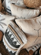 A collection of decorative pillows featuring intricate patterns and textures, some with black and white geometric designs and others with woven and embroidered details. The pillows are stacked closely together, creating a cozy and artisanal appearance. The materials include woven straw-like textures and soft fabric surfaces.