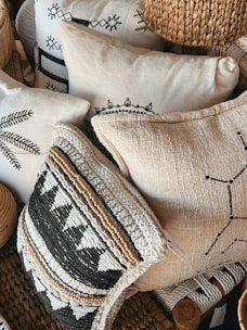 A collection of decorative pillows featuring intricate patterns and textures, some with black and white geometric designs and others with woven and embroidered details. The pillows are stacked closely together, creating a cozy and artisanal appearance. The materials include woven straw-like textures and soft fabric surfaces.