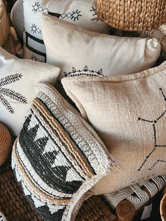 A collection of decorative pillows featuring intricate patterns and textures, some with black and white geometric designs and others with woven and embroidered details. The pillows are stacked closely together, creating a cozy and artisanal appearance. The materials include woven straw-like textures and soft fabric surfaces.