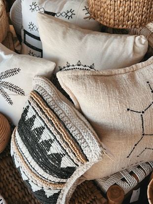 A collection of decorative pillows featuring intricate patterns and textures, some with black and white geometric designs and others with woven and embroidered details. The pillows are stacked closely together, creating a cozy and artisanal appearance. The materials include woven straw-like textures and soft fabric surfaces.