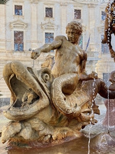 A detailed stone sculpture of a mythological scene with a figure riding a large fish-like creature, set against the background of a historic building. Water is splashing around, indicating a fountain setting.