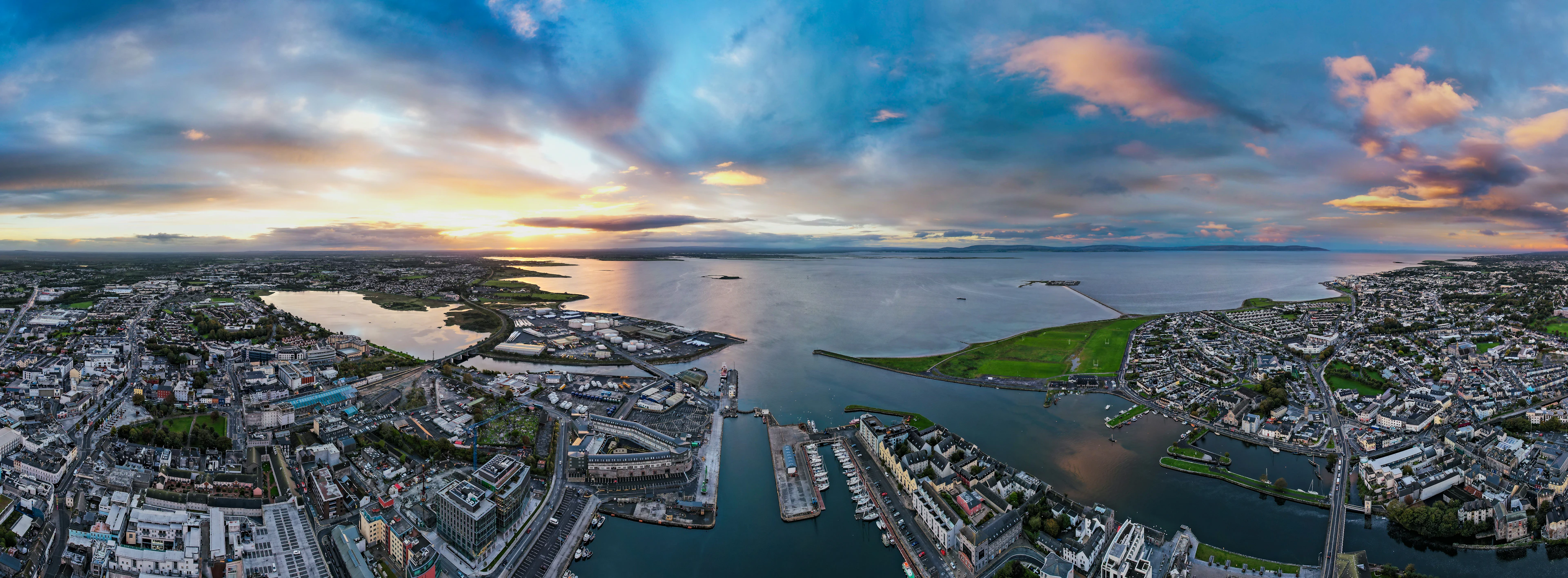 A wide aerial panorama of Galway at sunset, the River Corrib and the docks running out past the Claddagh into Galway Bay while the sun drops through broken cloud over the Atlantic