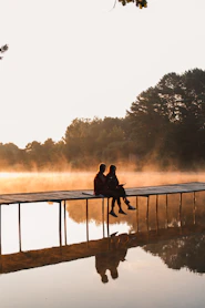 a man and woman sitting on a dock by a lake