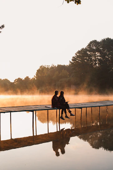 a man and woman sitting on a dock by a lake