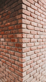 A close-up view of a brick corner wall constructed from reddish-brown bricks. The image captures the junction of two walls meeting at a right angle, showing the texture and alignment of bricks in a stacked formation.