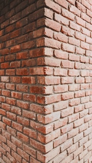 A close-up view of a brick corner wall constructed from reddish-brown bricks. The image captures the junction of two walls meeting at a right angle, showing the texture and alignment of bricks in a stacked formation.