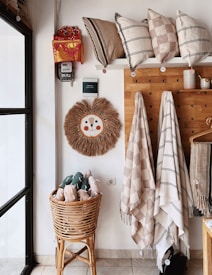 The image depicts an interior corner of a room with a cozy and decorative setup. There is a wicker basket filled with plush toys in the lower left, a wall-mounted pegboard holding various hanging textiles like towels and blankets, and multiple decorative pillows arranged on a shelf above. A unique round wall hanging with a textured, woven design featuring a smiling face is also noticeable. Additionally, a small shelf with decorative cups is attached to the pegboard.