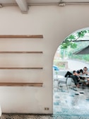 A minimalist interior with wooden shelves on a white wall is shown on the left, and an arched opening leads to a cozy outdoor seating area with patterned tile flooring. A group of people is gathered around a table, browsing their phones, while lush greenery is visible through the opening.