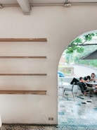 A minimalist interior with wooden shelves on a white wall is shown on the left, and an arched opening leads to a cozy outdoor seating area with patterned tile flooring. A group of people is gathered around a table, browsing their phones, while lush greenery is visible through the opening.
