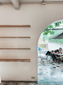 A minimalist interior with wooden shelves on a white wall is shown on the left, and an arched opening leads to a cozy outdoor seating area with patterned tile flooring. A group of people is gathered around a table, browsing their phones, while lush greenery is visible through the opening.