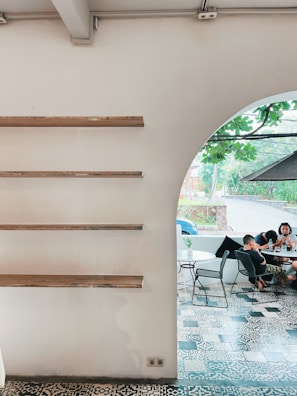 A minimalist interior with wooden shelves on a white wall is shown on the left, and an arched opening leads to a cozy outdoor seating area with patterned tile flooring. A group of people is gathered around a table, browsing their phones, while lush greenery is visible through the opening.