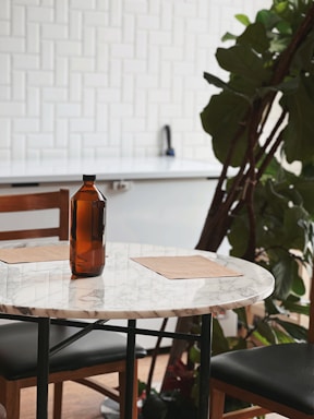 A round marble table with an amber glass bottle situated in a modern interior setting. Two placemats are placed on either side of the table, and a wooden chair with a dark cushion is partially visible. A large green plant is positioned nearby, and the background features a white tiled wall and a light-colored countertop.