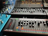 A collection of synthesizer modules with various knobs, sliders, and buttons arranged in an organized manner. The foreground features a device labeled 'JX-03' with two sliders glowing red and numerous knobs for controlling different parameters. Other synthesizers and modules are visible in the background, each with their own array of controls and LED indicators.