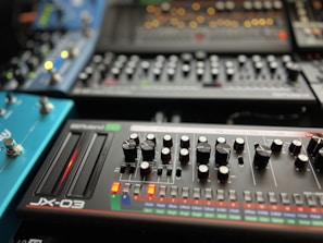 A collection of synthesizer modules with various knobs, sliders, and buttons arranged in an organized manner. The foreground features a device labeled 'JX-03' with two sliders glowing red and numerous knobs for controlling different parameters. Other synthesizers and modules are visible in the background, each with their own array of controls and LED indicators.