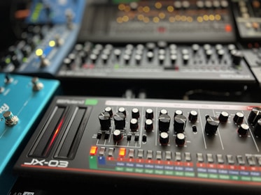 A collection of synthesizer modules with various knobs, sliders, and buttons arranged in an organized manner. The foreground features a device labeled 'JX-03' with two sliders glowing red and numerous knobs for controlling different parameters. Other synthesizers and modules are visible in the background, each with their own array of controls and LED indicators.
