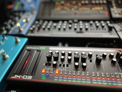 A collection of synthesizer modules with various knobs, sliders, and buttons arranged in an organized manner. The foreground features a device labeled 'JX-03' with two sliders glowing red and numerous knobs for controlling different parameters. Other synthesizers and modules are visible in the background, each with their own array of controls and LED indicators.