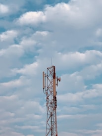 A tall communication tower with antennas and a satellite dish is set against a backdrop of a partly cloudy sky. The structure is composed of metal frameworks and various technological components.