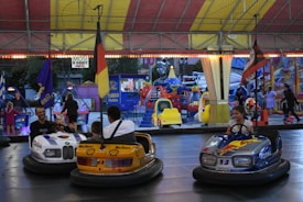 A lively amusement park scene with people enjoying a bumper car ride. A man and child are in a car designed like a police car, and another person is in a car with vibrant colors and branding like Marlboro and Shell. A young girl drives a blue bumper car with Total and Mobil logos. In the background, there are colorful rides and attractions, including a small ride shaped like helicopters. Flags and lights enhance the festive atmosphere.