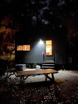 A small, dark cabin illuminated from the inside by warm lights. It is situated in a wooded area at night. The foreground features a wooden picnic table on gravel, with some foliage partially obscuring the view. A set of stairs leads to the cabin entrance, casting shadows across the scene.