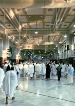 A large group of people dressed in white robes walk towards a destination in an intricately decorated building. The ceiling is adorned with ornate designs and light fixtures, while the walls feature decorative arches and columns. The atmosphere is solemn and reverent.