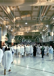 A large group of people dressed in white robes walk towards a destination in an intricately decorated building. The ceiling is adorned with ornate designs and light fixtures, while the walls feature decorative arches and columns. The atmosphere is solemn and reverent.