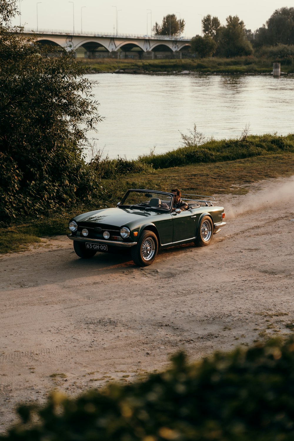 a car on a dirt road by a river with a bridge in the background