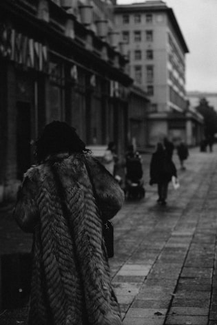A person wearing a fur coat walks down a city street, surrounded by buildings. The street is paved with stone tiles, and there are a few people in the distance, one pushing a stroller. The scene is in black and white, giving it a vintage or melancholic feel.