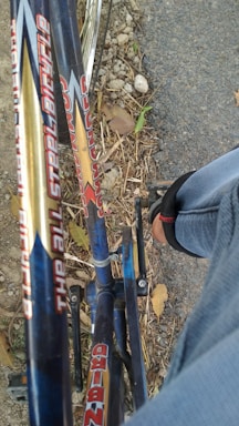 Close-up view of a bicycle's frame with the text 'The All Steel Bicycle' visible, parked on a road with some gravel and leaves scattered around. A person's leg wearing jeans and a sandal is partially visible, showing they are sitting on the bicycle.