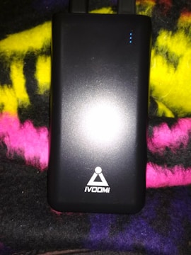 A black rectangular electronic device, likely a power bank, with the logo 'iVOOMi' printed on it. The device is placed on a multicolored fabric background with shades of yellow, pink, and purple. There are small blue indicator lights on the upper right corner of the device.