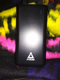 A black rectangular electronic device, likely a power bank, with the logo 'iVOOMi' printed on it. The device is placed on a multicolored fabric background with shades of yellow, pink, and purple. There are small blue indicator lights on the upper right corner of the device.