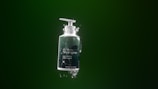 A bottle of shampoo with a pump dispenser is suspended in mid-air against a dark green background. The bottle is surrounded by splashes of water, creating a dynamic and fresh appearance. The label on the bottle reads 'Shampoo' and mentions 'Juicy fruits & warm woods', implying a refreshing and natural scent.