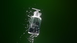 A bottle of shampoo with a pump dispenser is suspended against a dark green background. Water splashes dynamically around it, giving a fresh and clean impression. The design on the bottle includes subtle text and minimalistic graphics.