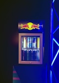 A compact beverage fridge with a transparent door filled with various cans of energy drinks. The fridge has a prominent logo on top featuring two red bulls and a yellow sun. The scene is lit with blue lighting, creating a vibrant and dynamic atmosphere.