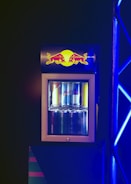 A compact beverage fridge with a transparent door filled with various cans of energy drinks. The fridge has a prominent logo on top featuring two red bulls and a yellow sun. The scene is lit with blue lighting, creating a vibrant and dynamic atmosphere.