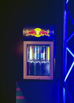 A compact beverage fridge with a transparent door filled with various cans of energy drinks. The fridge has a prominent logo on top featuring two red bulls and a yellow sun. The scene is lit with blue lighting, creating a vibrant and dynamic atmosphere.