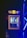 A compact beverage fridge with a transparent door filled with various cans of energy drinks. The fridge has a prominent logo on top featuring two red bulls and a yellow sun. The scene is lit with blue lighting, creating a vibrant and dynamic atmosphere.
