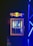A compact beverage fridge with a transparent door filled with various cans of energy drinks. The fridge has a prominent logo on top featuring two red bulls and a yellow sun. The scene is lit with blue lighting, creating a vibrant and dynamic atmosphere.