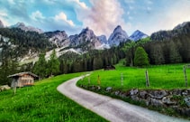 A picturesque landscape featuring a winding path leading through lush green meadows towards rugged mountain peaks. Two rustic wooden huts can be seen among the vibrant greenery, while dense forests cover the hills. Majestic mountains loom in the background under a partly cloudy blue sky.