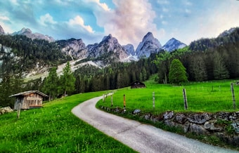 A picturesque landscape featuring a winding path leading through lush green meadows towards rugged mountain peaks. Two rustic wooden huts can be seen among the vibrant greenery, while dense forests cover the hills. Majestic mountains loom in the background under a partly cloudy blue sky.