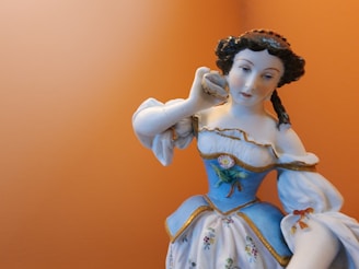 A porcelain figurine of a woman with dark braided hair, dressed in a blue and white gown, is holding a shell to her ear. The background is a soft orange color.