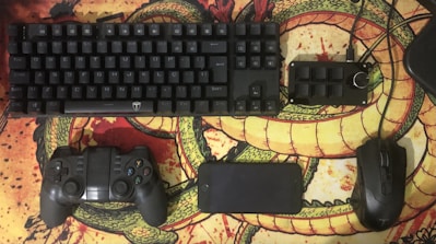 A computer keyboard, game controller, smartphone, additional keypad, and a computer mouse are arranged neatly on a desk mat featuring a dragon design. The items are predominantly black and contrast with the vibrant, colorful background.