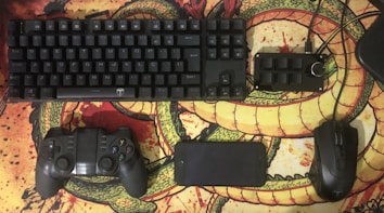 A computer keyboard, game controller, smartphone, additional keypad, and a computer mouse are arranged neatly on a desk mat featuring a dragon design. The items are predominantly black and contrast with the vibrant, colorful background.