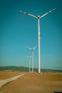 Three wind turbines stand in an open landscape with clear skies above. The turbines are aligned along a dirt path that cuts through the vast, flat expanse of earth-toned fields. Hills with patches of greenery can be seen in the distance.