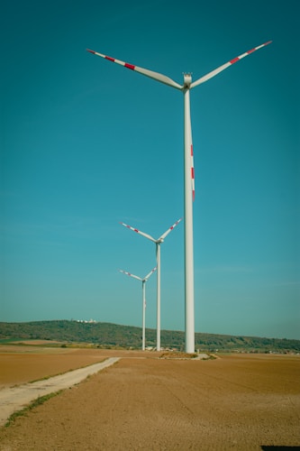 Three wind turbines stand in an open landscape with clear skies above. The turbines are aligned along a dirt path that cuts through the vast, flat expanse of earth-toned fields. Hills with patches of greenery can be seen in the distance.